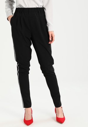 Femme portant un pantalon noir slim avec des bandes blanches sur les côtés, une chemise blanche à manches longues et des talons hauts rouges pointus, debout avec une main dans la poche.