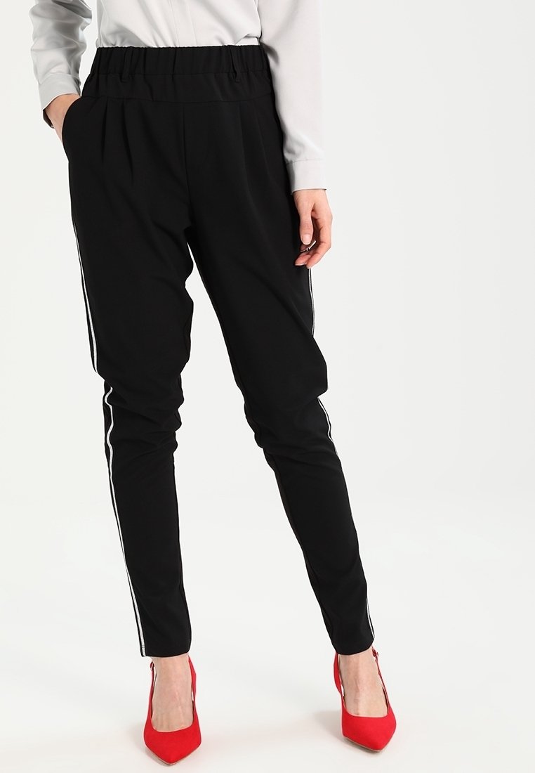 Femme portant un pantalon noir slim avec des bandes blanches sur les côtés, une chemise blanche à manches longues et des talons hauts rouges pointus, debout avec une main dans la poche.