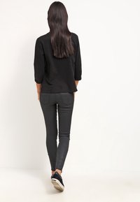 Top negro de manga tres cuartos, combinado con pantalones negros ajustados y zapatillas oscuras, visto desde atrás sobre un fondo blanco liso.