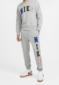 Šedá mikina se vícebarevným logem "NIKE"; odpovídající tepláky s nápisem "NIKE" na noze; texturední látka; bílé tenisky s červenými akcenty.
