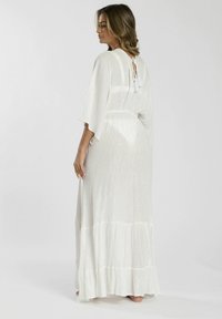 Largo vestido blanco con una textura brillante, mangas sueltas y un dobladillo en capas. Los lazos en la parte posterior cuentan con borlas decorativas.