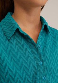 Cămașă teal cu guler, având un model texturat herringbone, butoane albastre și o croială lejeră dintr-un material moale.
