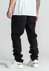 Pantalones de pana negros con un corte holgado, con bolsillos traseros y combinados con zapatillas blancas que muestran un logo de la marca en el lateral.