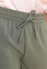 Joggings à taille élastique en tissu vert olive, présentant un détail à nouer et une texture lisse avec un léger éclat. Gros plan sur la zone de la taille.