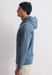 Blauer Kapuzenpullover mit durchgehendem Reißverschluss vorne, verstellbarer Kapuze und Seitentaschen. Wird über hellen Hosen getragen und mit einer strukturierten dunklen Mütze kombiniert.