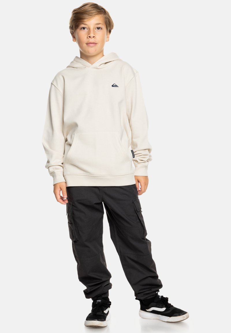 Beige hoodie met voorzak, gecombineerd met zwarte cargobroek; model draagt zwart-witte sneakers. Eenvoudig ontwerp, casual pasvorm.