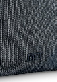Tejido negro texturizado con un patrón sutil; presenta un logo oscuro en relieve "JOST" cerca de la esquina inferior. Forma redondeada.