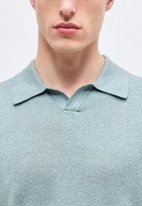 Κοντινό πλάνο ενός ατόμου που φοράει ανοιχτό μπλε πλεκτό πόλο μπλουζάκι με απαλό γιακά και μικρό άνοιγμα σε σχήμα V.