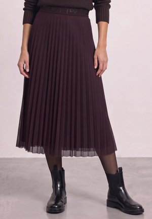 Le mannequin porte une jupe midi plissée couleur prune foncé avec un pull assorti, un collant noir et des bottines noires.