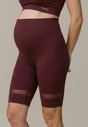 Femme portant un short de sport bordeaux taille haute et un crop top assorti avec un ventre de grossesse visible, debout sur un fond uni.