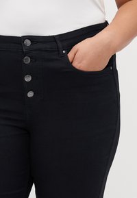 Jeans de mezclilla negra de tiro alto, con una textura suave, que cuentan con un cierre frontal de cinco botones y bolsillos laterales.