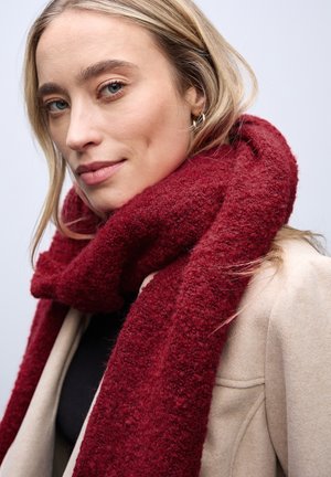 Écharpe en laine bourgogne avec une finition texturée et douce, enroulée autour du cou ; portée avec un manteau beige clair et un haut noir en dessous.