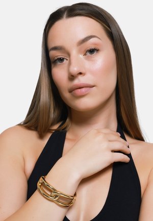 Bracciale in metallo dorato con design a catena intrecciata. Finitura liscia e lucida con una forma elegante e moderna. Indossato su un braccio nudo.