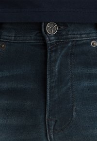 Tamno plave traperice s klasičnim ravnim krojem, metalnim dugmetom za zatvaranje i suptilnom teksturiranom završnom obradom. Sadrže kontrastno šivanje i džepove.