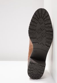 Semelle de botte marron avec des motifs de bande de roulement profonds, comprenant des dessins en forme de fleur, présentée verticalement sur un fond blanc.