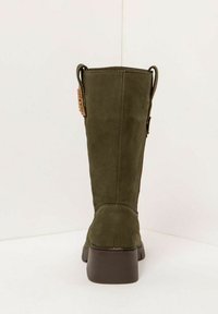Bota alta de ante verde oliva con tacón cuadrado marrón y pestañas de agarre, mostrada desde la parte posterior contra un fondo blanco liso.