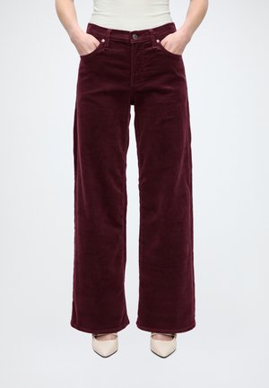 Un mannequin porte un pantalon bordeaux à jambes larges, un haut blanc et des chaussures pointues couleur crème, les mains dans les poches.
