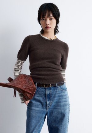 Brown knit sweater layered over a striped long-sleeve shirt, paired with blue jeans and holding a woven brown handbag. Gold chain necklace accent.