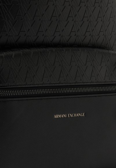 Gros plan sur un sac noir Armani Exchange avec des lettres géométriques gaufrées, une fermeture zippée et un petit logo métallique.
