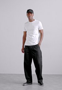 T-shirt blanche à manches courtes avec un petit logo, associée à un pantalon noir ample et des baskets noires. La casquette est également noire.