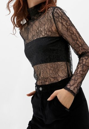 Woman wearing a fitted black lace long-sleeve top over a black bandeau, paired with high-waisted black pants, hands in pockets.