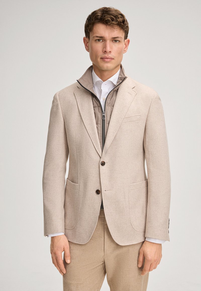 Jasnobeżowa, dopasowana marynarka z dwoma przednimi kieszeniami i brązowymi guzikami, noszona na jasnej, ocieplanej kamizelce. Pod spodem biała koszula wizytowa.