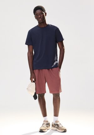 Mladý muž v námorníckom modrom tričku, hrdzavo sfarbených šortkách, béžových teniskách a držiaci čiernu a béžovú čiapku na jednoliatom bielom pozadí.