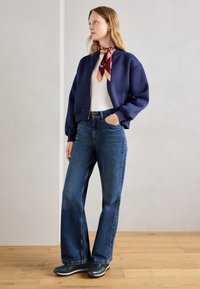 Bombardier bleu marine, haut léger, jeans bleus taille haute, stylés avec une écharpe à motifs. Chaussures noires avec semelle striée, fond en parquet.
