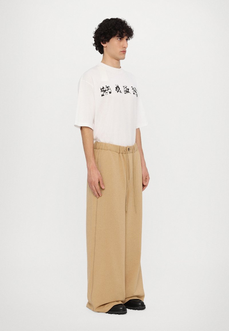 Weißes oversized T-Shirt mit schwarzem Grafikdruck, kombiniert mit beigen weit geschnittenen Hosen. Die Hosen haben einen Kordelzugbund und eine weiche Textur.