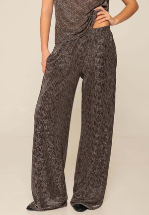 Mujer vistiendo pantalones sueltos marrones de pierna ancha con textura y top a juego, de pie con una mano en la cadera contra un fondo neutro.