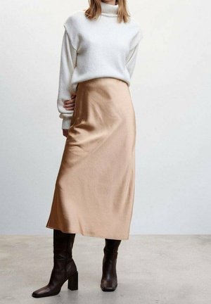 Gonna lunga in satin beige con una texture liscia, abbinata a un maglione bianco largo e stivali marroni con tacco fino al ginocchio.