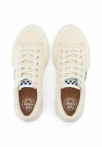 Zapatillas de lona beige con cordones blancos, diseño de punta redonda y plantilla de corcho texturizada. Presenta una etiqueta a rayas en blanco y negro en el costado.