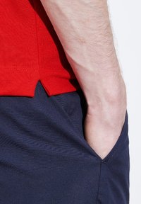 Röd polo skjorta med en texturerad tyg, med en spjälld nederkant. Mörkblå byxor med sidficka, som har en slät finish.