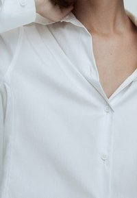 Chemise blanche à boutons avec un col en V, manches longues et tissu lisse. Comprend des boutons discrets et une coupe ajustée.
