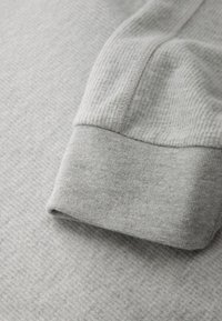 Graues Langarmshirt mit einer strukturierten, gerippten Stoffstruktur und einem Ärmelabschluss mit Manschette. Das Material wirkt weich und leicht, ideal zum Unterziehen.