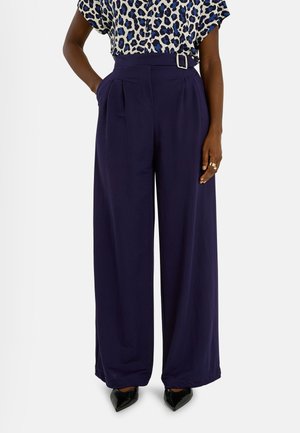 Mujer con pantalones anchos azul marino con hebilla en el cinturón, combinados con blusa de estampado de leopardo y tacones negros de punta afilada, manos en los bolsillos.