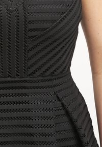 Robe noire sans manches avec des rayures en maille transparente alternant entre horizontales et diagonales, taille cintrée et un détail peplum sur le côté.
