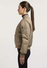 Khaki Bomberjacke aus einem glatten, leichten Material. Verfügt über eine Reißverschlusstasche am Arm und eine Frontklapptasche, mit gerippten Bündchen und Saum.