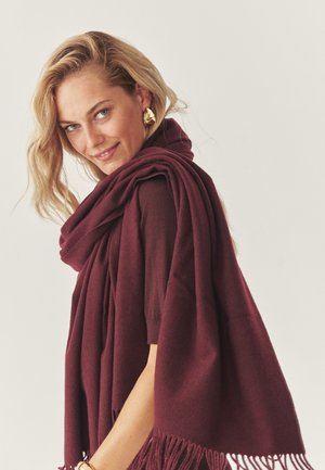 Burgundská šatka s rozstrapkanými okrajmi, vyrobená z mäkkého materiálu, prehodená cez plecia. Modelove tričko má tmavý odtieň, ktorý dopĺňa šatku.