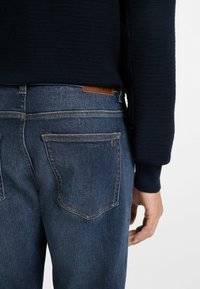 Jeans de mezclilla en azul oscuro con una textura suave y costuras naranjas en contraste. Incluye dos bolsillos traseros y un parche de logo de cuero.