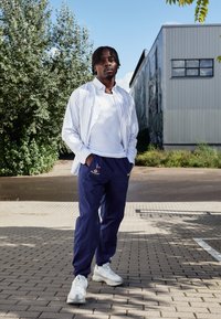 Weißes, gestreiftes Hemd über einem weißen Tanktop getragen, kombiniert mit marineblauen Hosen mit einem bunten Logo und weißen Sneakers.