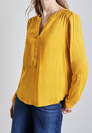 Femme portant un chemisier jaune moutarde à manches longues avec patte de boutonnage devant et un jean bleu sur un fond clair uni.