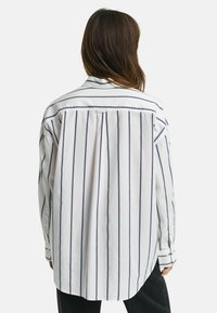 Witte shirt met verticale marineblauwe strepen, met knoopsluiting op de kraag en lange mouwen. Heeft een relaxte pasvorm en een geplisseerde achterkant.