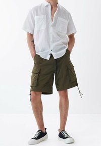 Uomo che indossa una camicia bianca a maniche corte con bottoni, pantaloni cargo verde oliva e sneaker nere a bassa forma con suola e lacci bianchi.