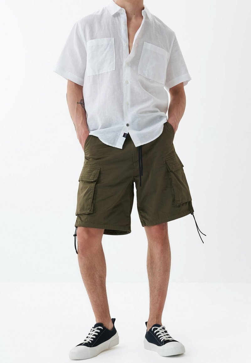 Uomo che indossa una camicia bianca a maniche corte con bottoni, pantaloni cargo verde oliva e sneaker nere a bassa forma con suola e lacci bianchi.