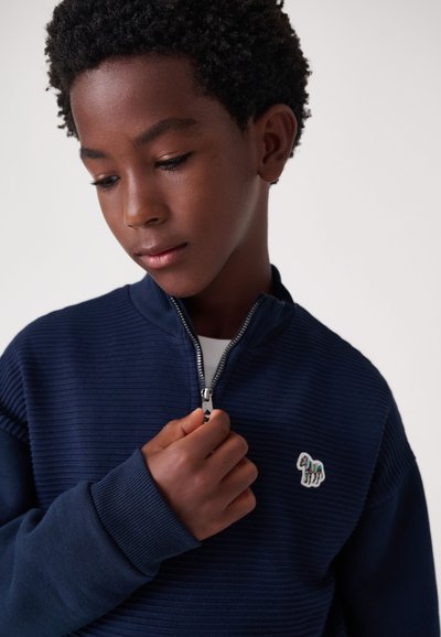 Tumši zils ribots džemperis ar rāvējslēdzēju un augstu apkakli, uz krūtīm izšūts dzīvnieka logo. Modelis tur rāvējslēdzēju ar vienu roku.