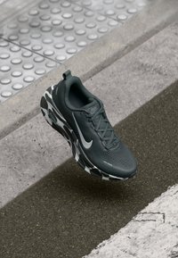 Tmavošedé bežecké topánky Nike s podrážkou v maskáčovom vzore sa vznášajú nad textúrovaným chodníkom a dopravnými značkami.