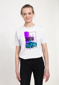 Hvid t-shirt med et grafisk print, der viser en blå bil, lilla malerispidser og teksten "TOKYO SPEED SHOP". Slim fit sorte bukser bæres.