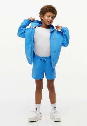 Niño con sudadera con capucha azul claro con cremallera y pantalones cortos a juego, camiseta blanca, calcetines blancos y zapatillas blancas, de pie sobre un fondo blanco.