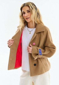 Žena s blond vlasmi oblečená v béžovom vlnenom kabáte, ktorý ukazuje jeho jasne červenú podšívku, bielu košeľu, krémové nohavice a vrstvené náhrdelníky.